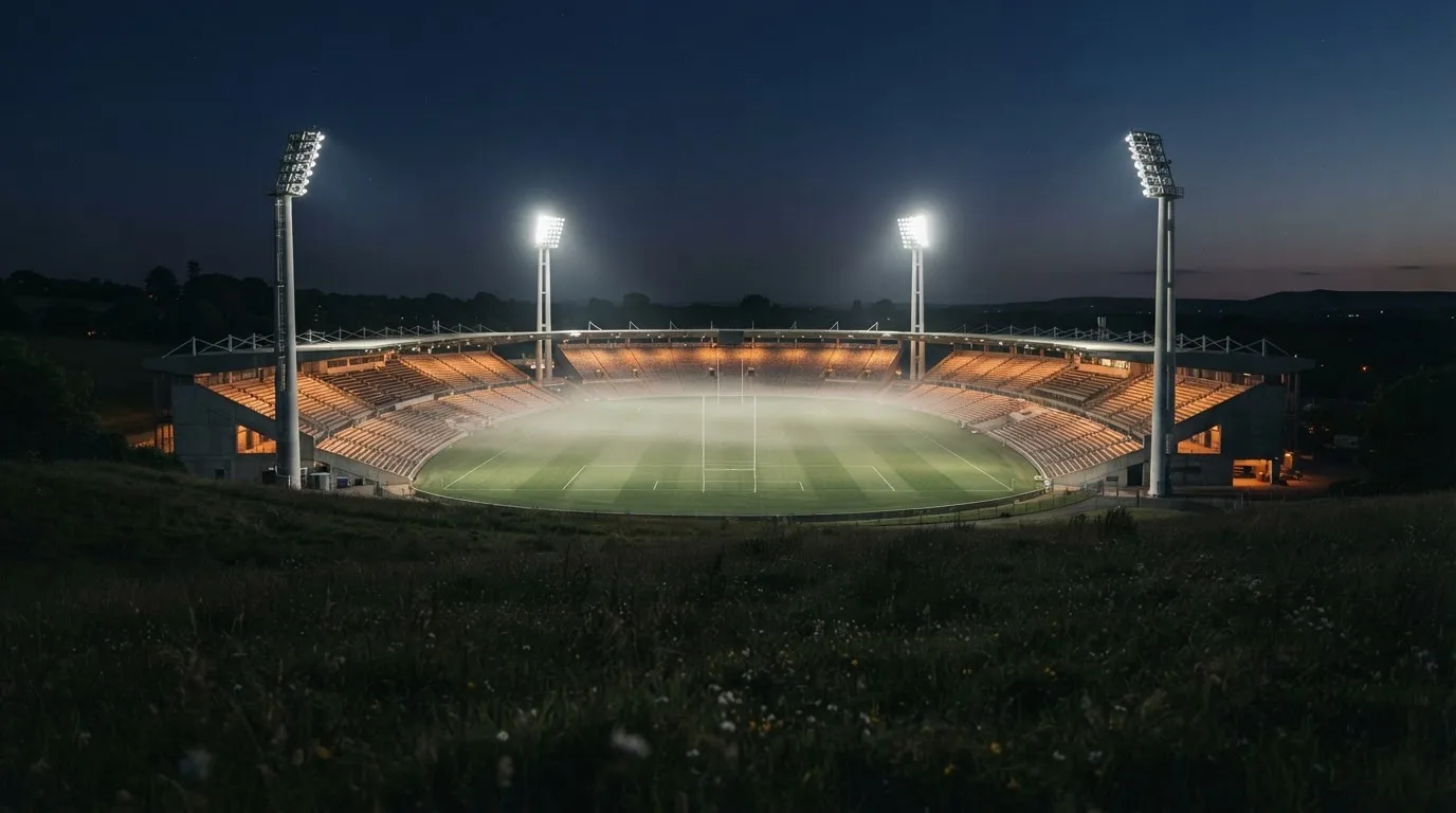 Ein Stadion unter Flutlicht bei Nacht, von einem Hügel aus der Ferne betrachtet, mit Dunst über dem Spielfeld.