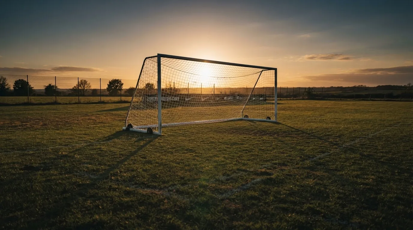 Ein Fußballtor mit Netz bei Sonnenuntergang auf einem leeren Platz, die Schatten der Torpfosten liegen lang auf dem Gras.