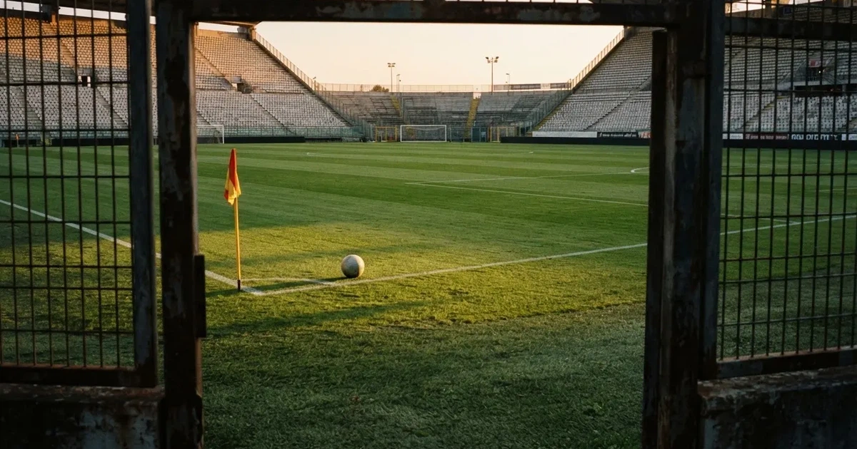 Blick durch ein Stadiontor auf einen grünen Rasen im Abendlicht, ein Ball ruht an der Eckfahne, die Tribünen sind leer.