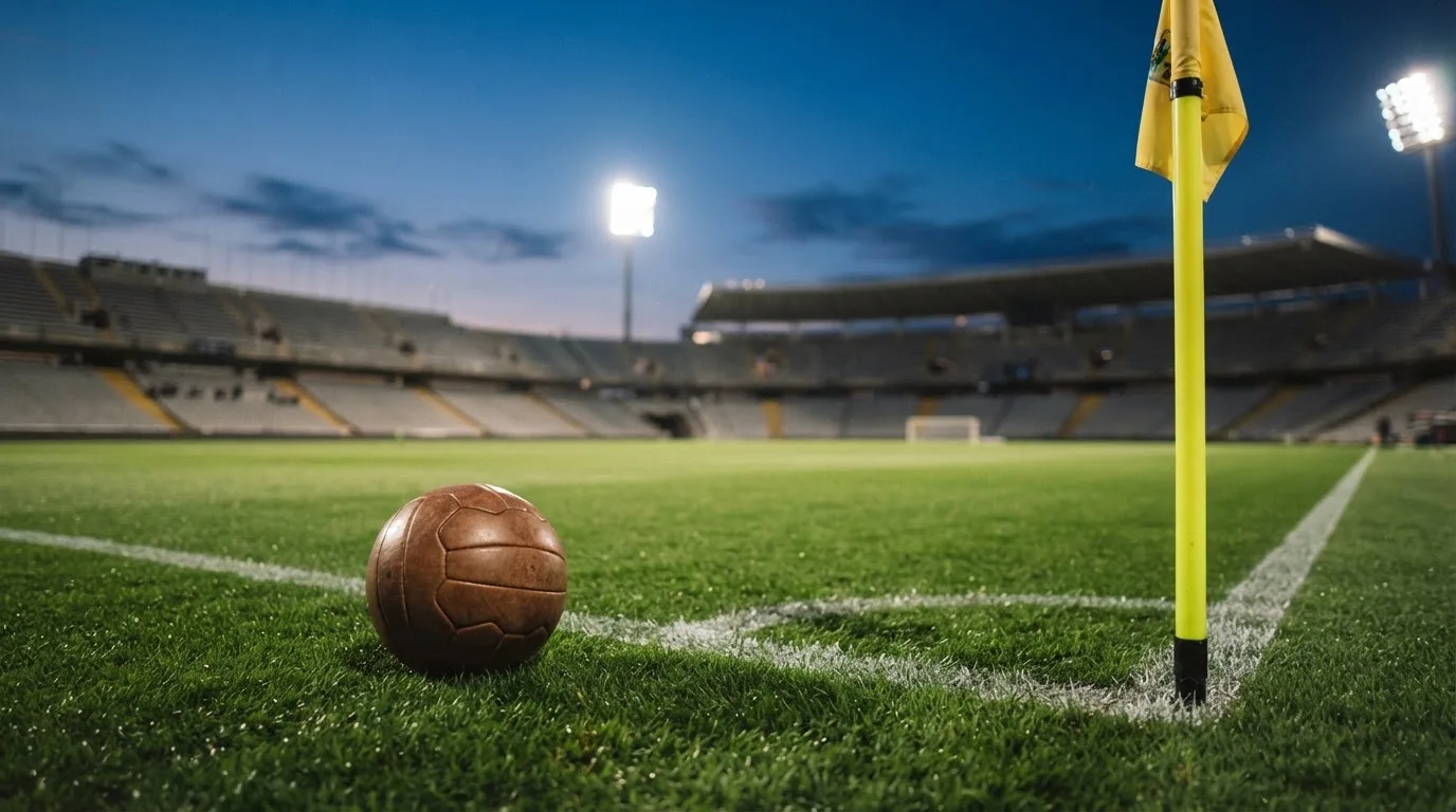 Ein Fußball liegt neben einem Eckfahnen-Pfosten auf einem Rasenplatz, im Hintergrund ein leeres Stadion in der Dämmerung.