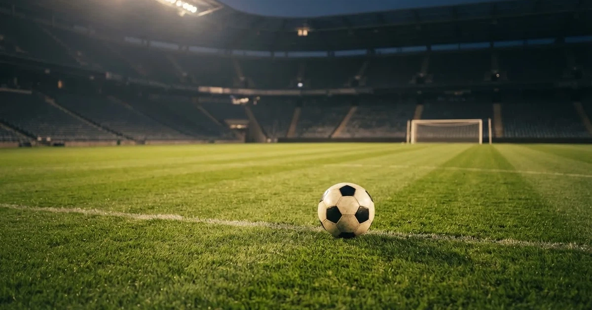 Ein Fußball liegt auf dem Rasen eines leeren Stadions bei Flutlicht, im Hintergrund unscharfe Tribünen ohne erkennbare Beschriftung.