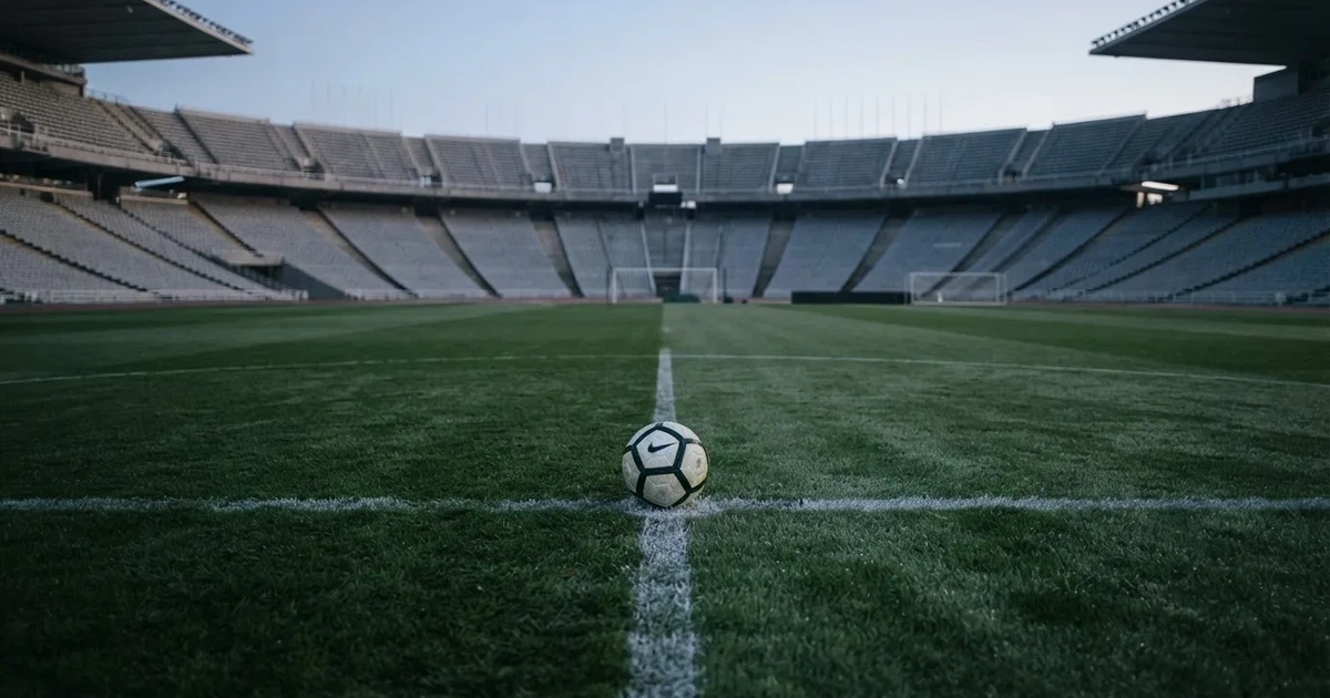 Ein Fußball liegt auf dem Mittelkreis eines Stadionrasens am frühen Morgen, im Hintergrund leere Ränge ohne erkennbare Beschriftung.