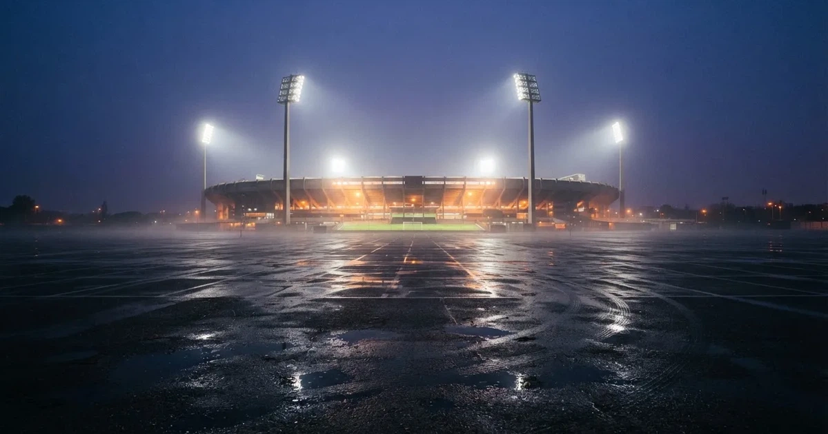 Ein Stadion unter Flutlicht aus großer Entfernung gesehen, davor ein regennasser Parkplatz in der Dämmerung.