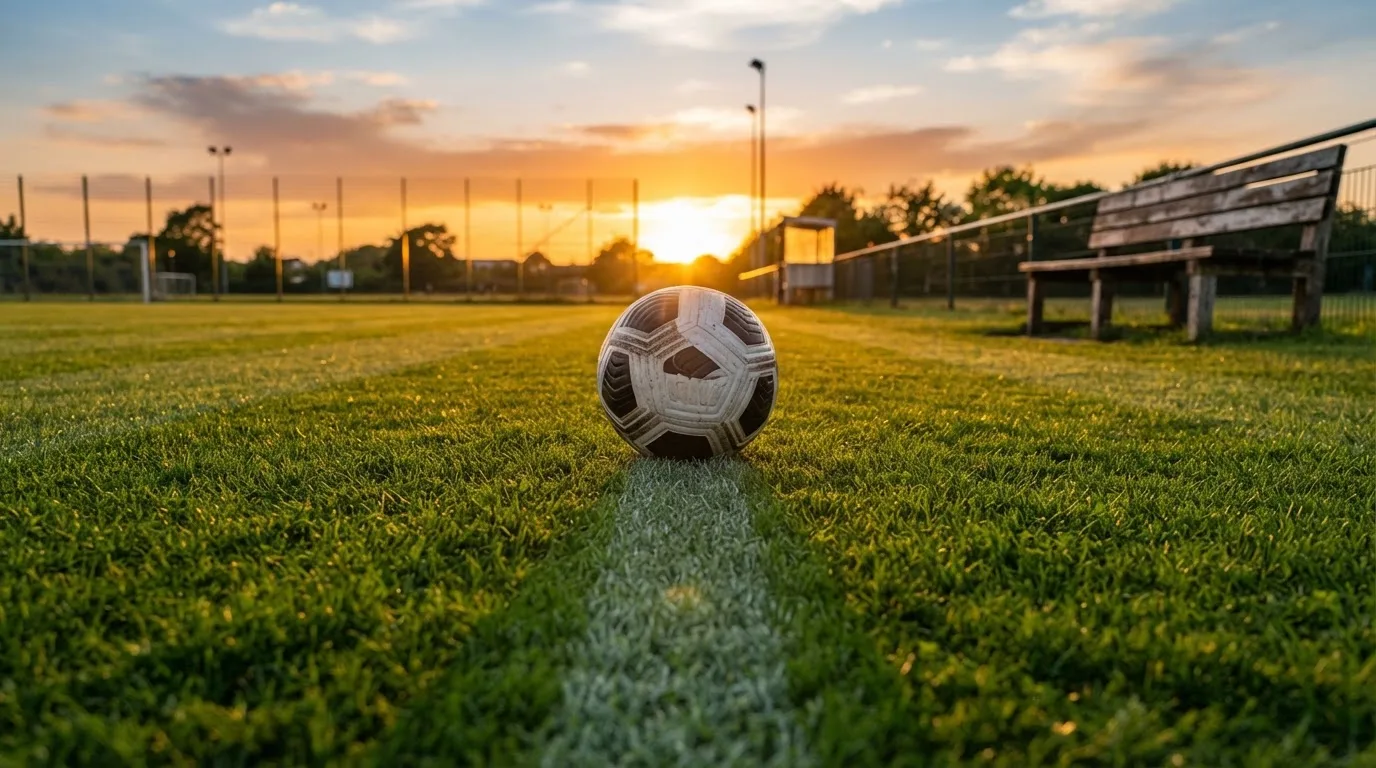 Ein Fußball auf einem Rasenplatz im Gegenlicht der tiefstehenden Sonne, am Spielfeldrand eine leere Ersatzbank.
