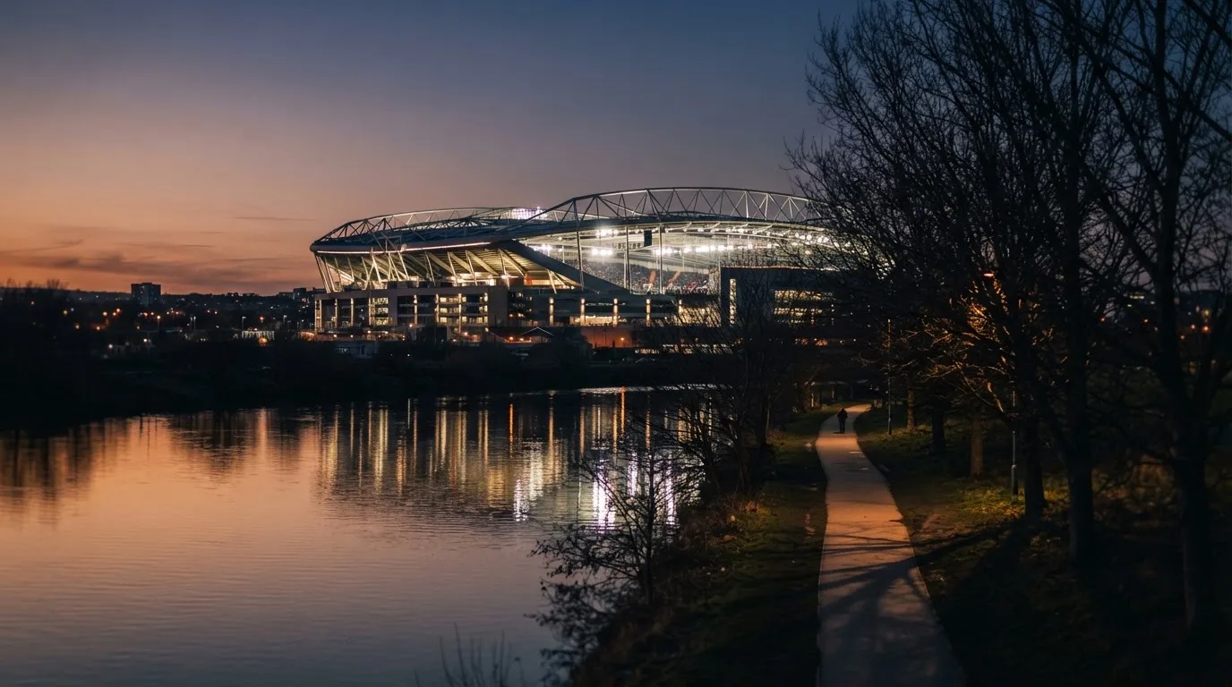 Ein Stadion unter Flutlicht aus der Ferne gesehen, im Vordergrund ein ruhiger Fluss und ein Fußweg.