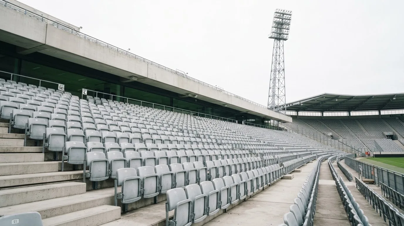Eine leere Tribüne eines Fußballstadions bei Tageslicht, Sitzschalen in Reihen und dahinter ein Flutlichtmast.