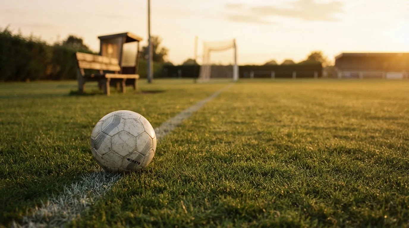 Ein Fußball liegt im Gras am Spielfeldrand, dahinter eine leere Bank und ein Tor im Gegenlicht.