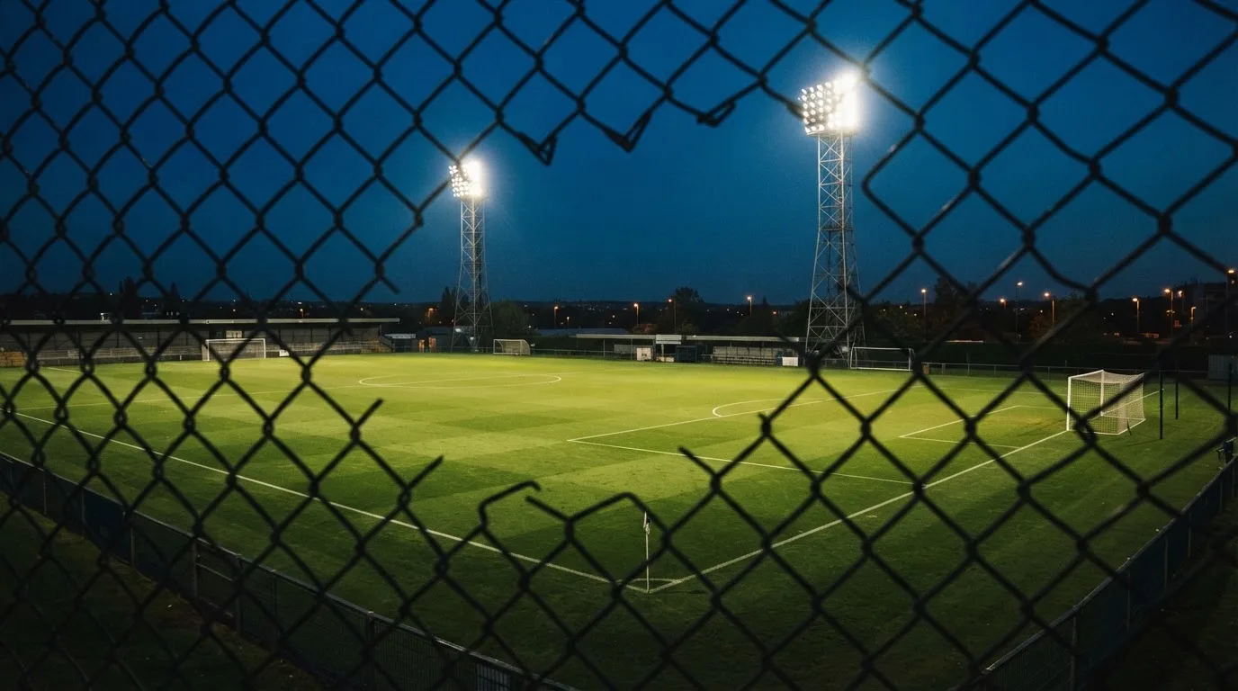 Eine Flutlichtanlage über einem leeren Sportplatz am Abend, im Vordergrund ein Maschendrahtzaun.