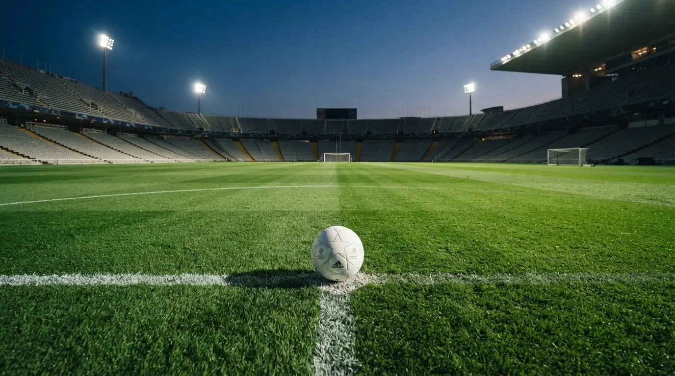 Ein Fußball liegt auf dem Anstoßpunkt eines leeren Rasenplatzes, im Hintergrund ein Stadion unter Flutlicht in der Abenddämmerung.