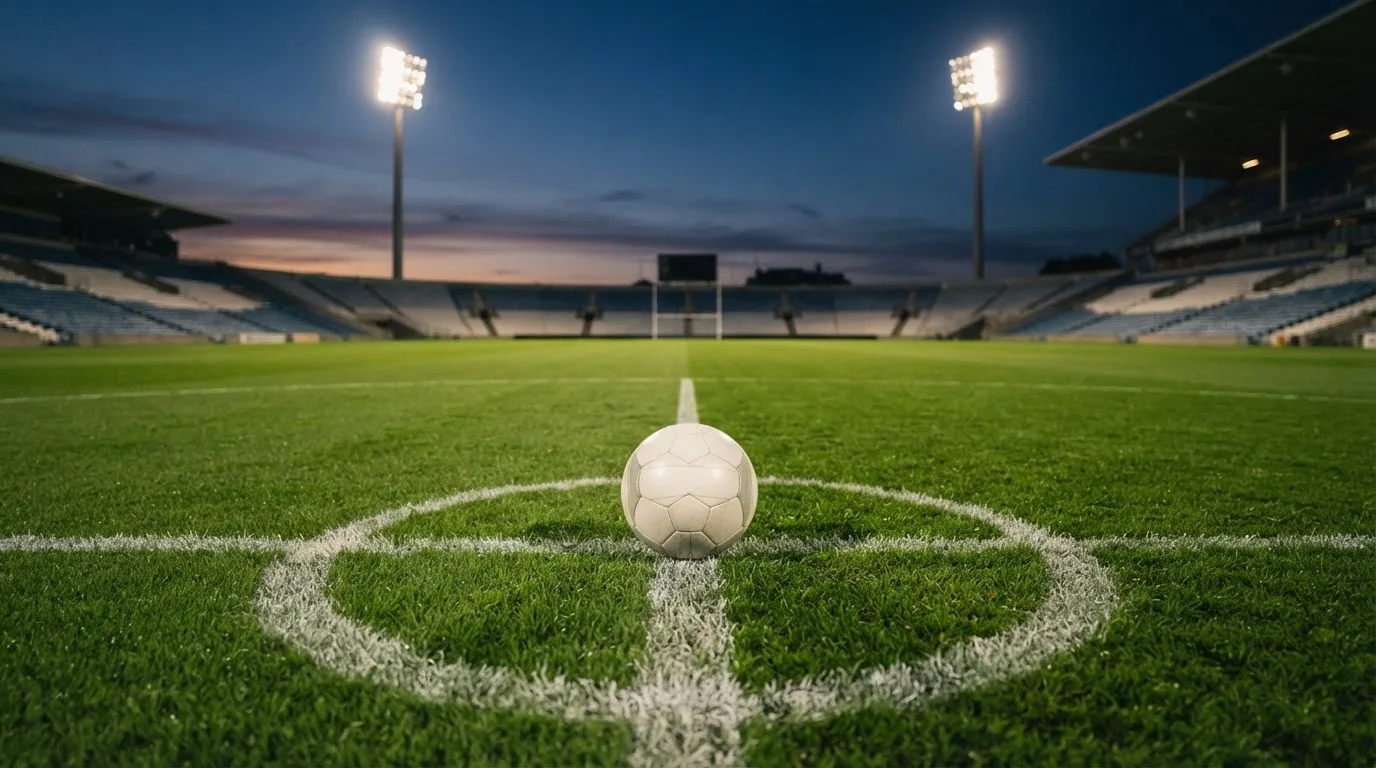 Ein Fußball liegt auf dem Anstoßpunkt eines leeren Rasenplatzes, im Hintergrund steht ein Stadion unter Flutlicht in der Dämmerung.