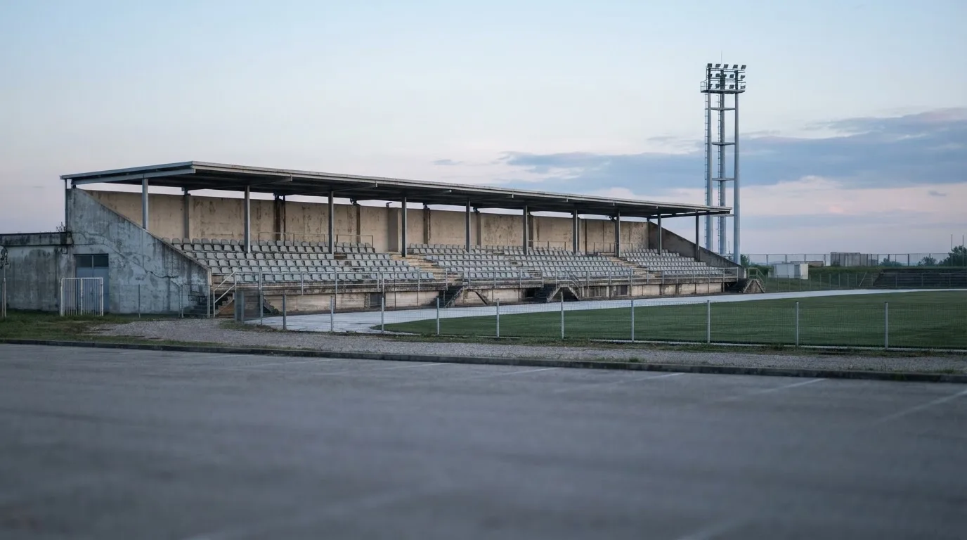 Eine leere Tribüne eines kleinen Stadions am frühen Morgen, aus der Ferne über einen menschenleeren Parkplatz fotografiert.