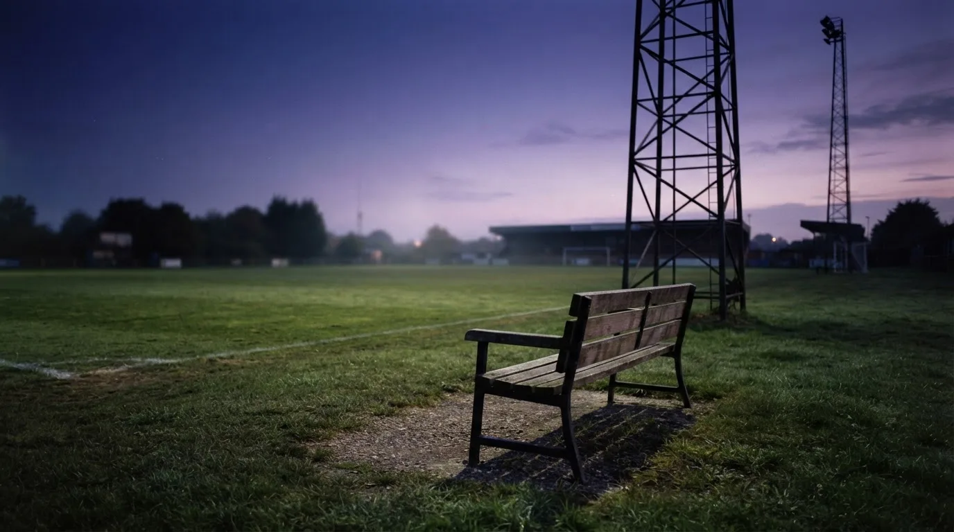 Eine leere Parkbank am Rand eines Fußballfelds in der Abenddämmerung, im Hintergrund ragen dunkle Flutlichtmasten in den Himmel.