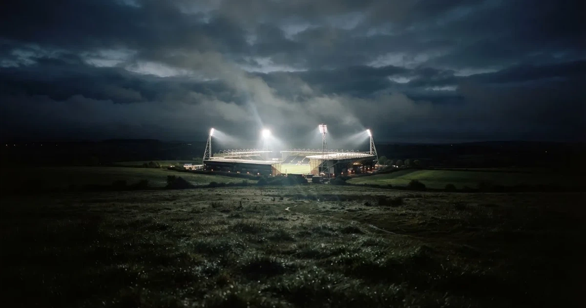 Ein Stadion unter Flutlicht, aus großer Entfernung über eine dunkle Wiese fotografiert, am Himmel zieht Nebel auf.