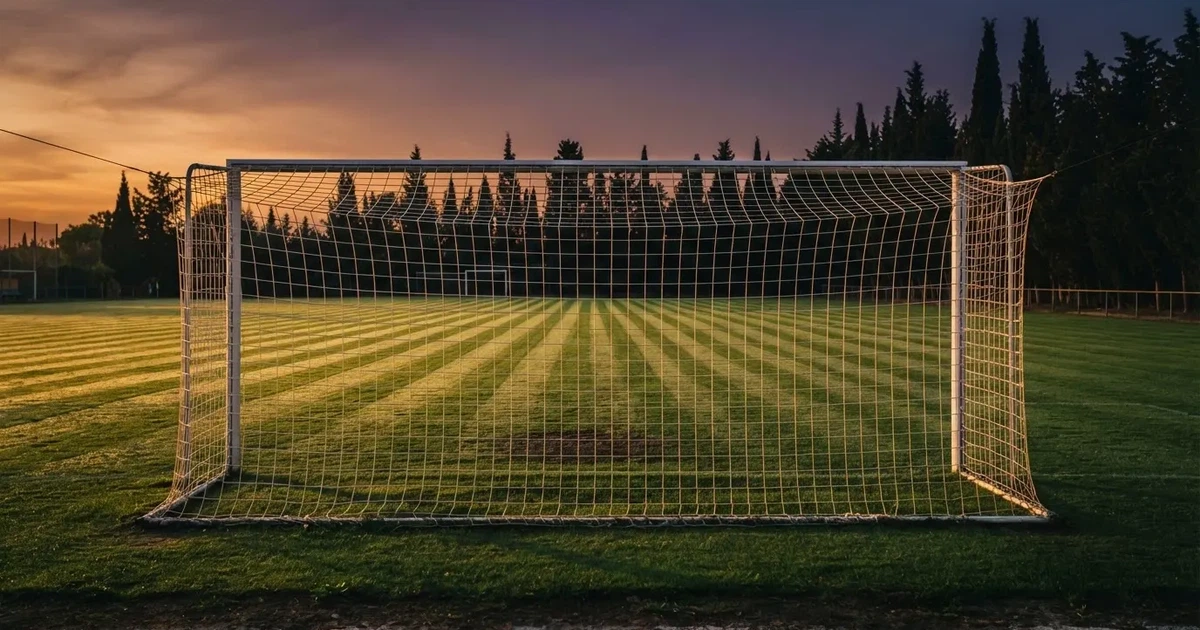 Ein Fußballtor mit Netz auf einem leeren Platz im Abendlicht, der Rasen frisch gemäht, im Hintergrund Zypressen.