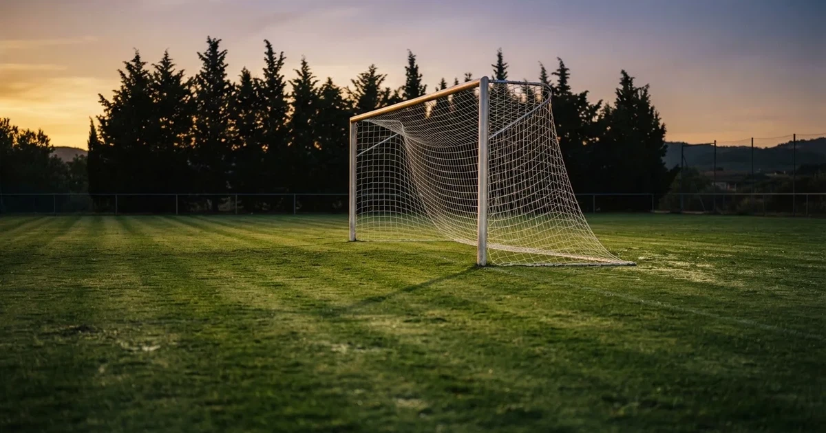 Ein Fußballtor mit Netz auf einem leeren Platz im Abendlicht, der Rasen frisch gemäht, im Hintergrund Zypressen.