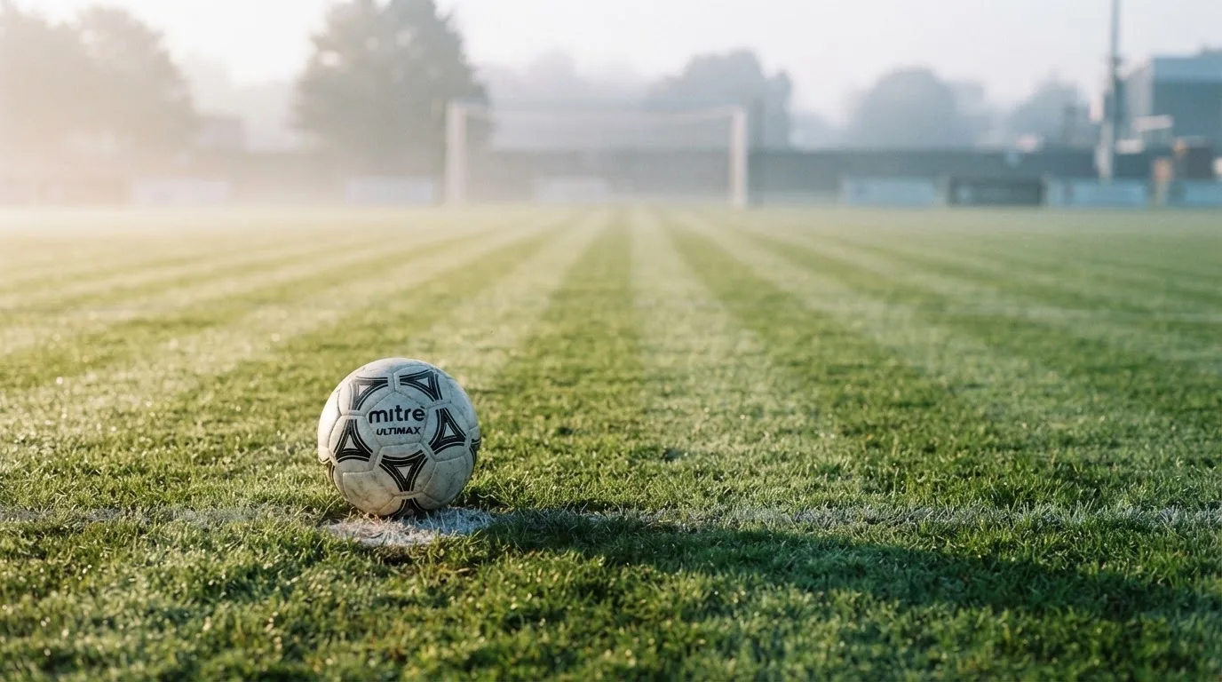 Ein Fußball liegt auf dem Elfmeterpunkt eines leeren Rasenplatzes, im Hintergrund unscharf ein Tor im Morgendunst.