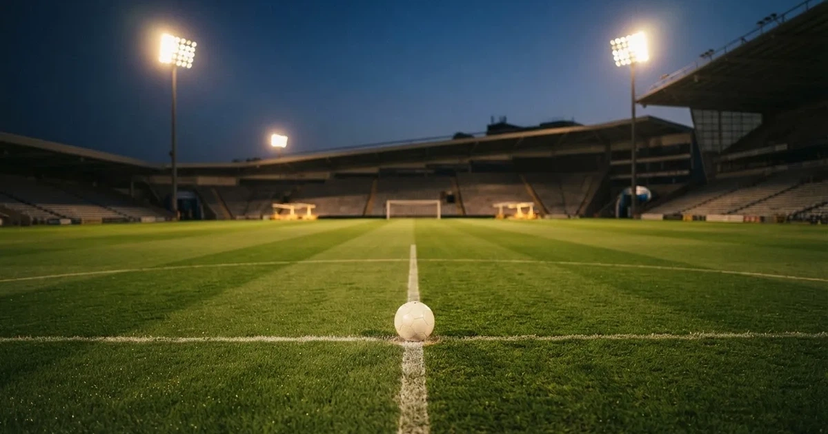Ein Rasenplatz in der Dämmerung mit eingeschalteten Flutlichtmasten, ein Ball liegt am Anstoßpunkt, die Tribünen im Hintergrund sind unscharf.