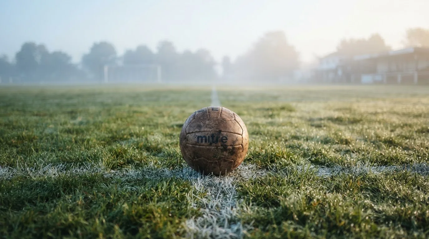 Ein abgewetzter Fußball liegt auf dem Anstoßkreis eines Naturrasens im Morgennebel.