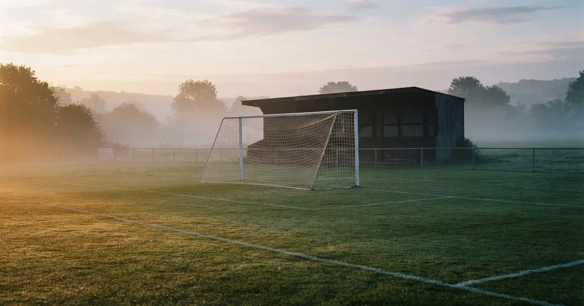 Ein Fußballtor mit Netz steht im Morgennebel auf einem leeren Rasenplatz, im Hintergrund die Silhouette einer kleinen Tribüne.
