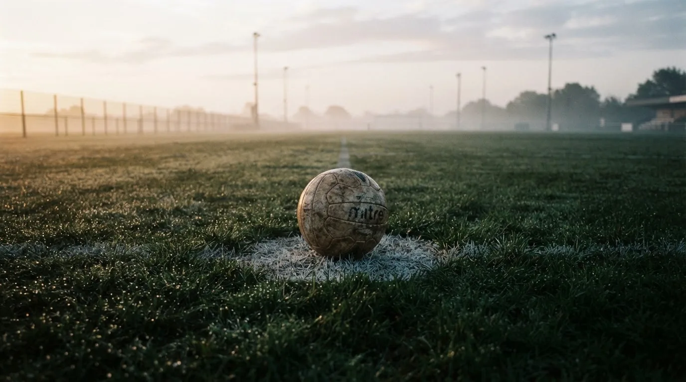 Ein Fußball liegt allein auf dem Anstoßpunkt eines leeren Rasenplatzes im Morgenlicht, am Rand ein niedriger Zaun.