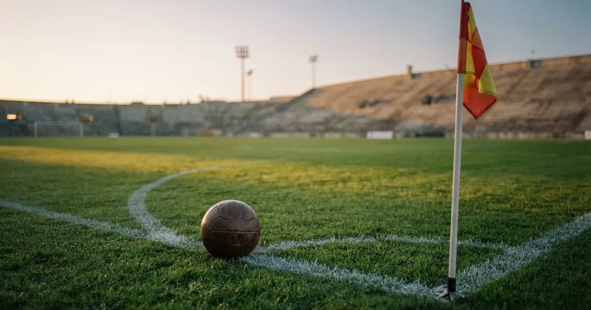 Ein Fußball liegt am Viertelkreis an der Eckfahne eines Rasenplatzes, im Hintergrund leere Tribünen in warmem Abendlicht.