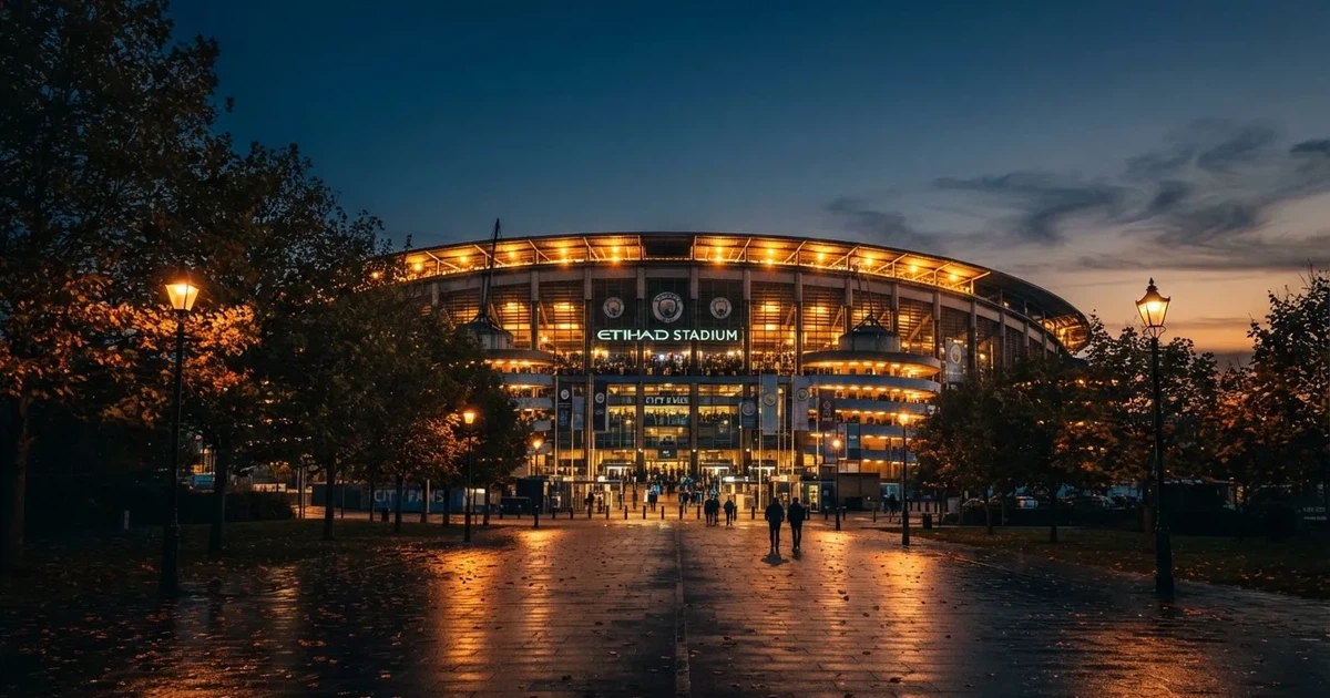 Ein großes Fußballstadion mit hell erleuchtetem Rund, aus der Ferne am Abend gesehen, davor eine breite Allee mit Laternen.